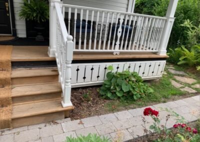 Garden bed beside wooden porch steps and a white railing, with green foliage and a red flower near the walkway.